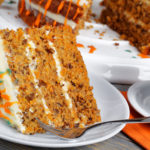 piece of classic carrot cake with cream cheese frosting decorated with walnuts and drizzled with colorful ganache with cup of tea on wooden table, view from above, close-up